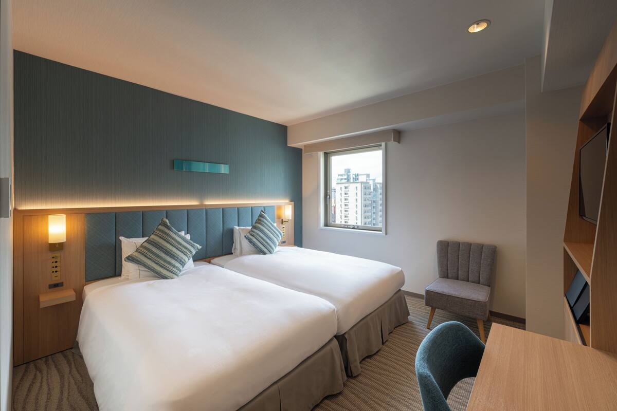 A hotel room with two beds, a small table, a chair, a TV, and a window with a city view.