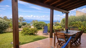 Eine überdachte Terrasse mit Tisch und Stühlen, ein blauer Liegestuhl und Blick auf das Meer und die Hügel.