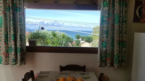 Ein Esstisch mit Obstschale, Holzstühle und Blick auf das Meer durch ein Fenster mit gemusterten Vorhängen.