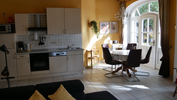 Une cuisine avec des meubles blancs, une table à manger avec des chaises et un canapé noir avec des coussins jaunes.