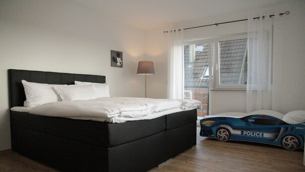 Un dormitorio con una cama negra, ropa de cama blanca, una lámpara, una ventana con cortinas y un coche de juguete de policía.