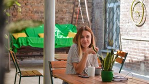 Une personne est assise à une table en bois, devant une plante verte, une tasse blanche et un téléphone intelligent.
