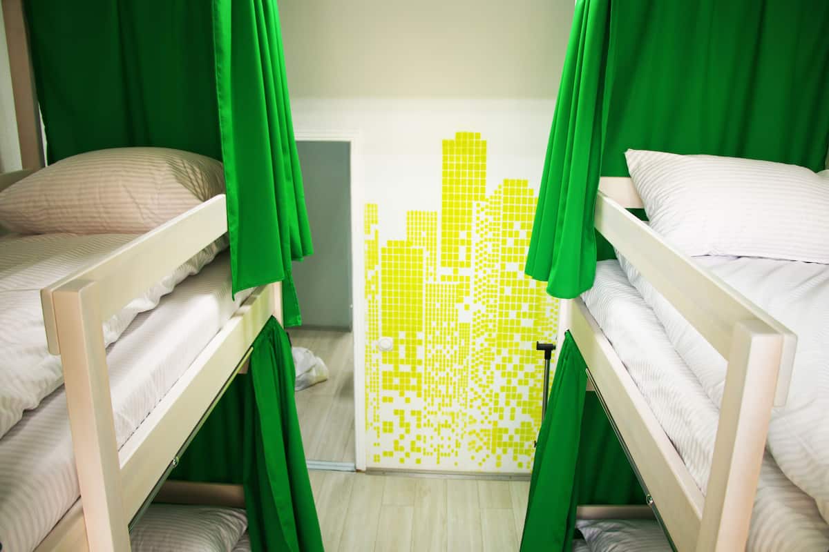 Una habitación con literas, cortinas verdes y un mural pixelado de un paisaje urbano amarillo.