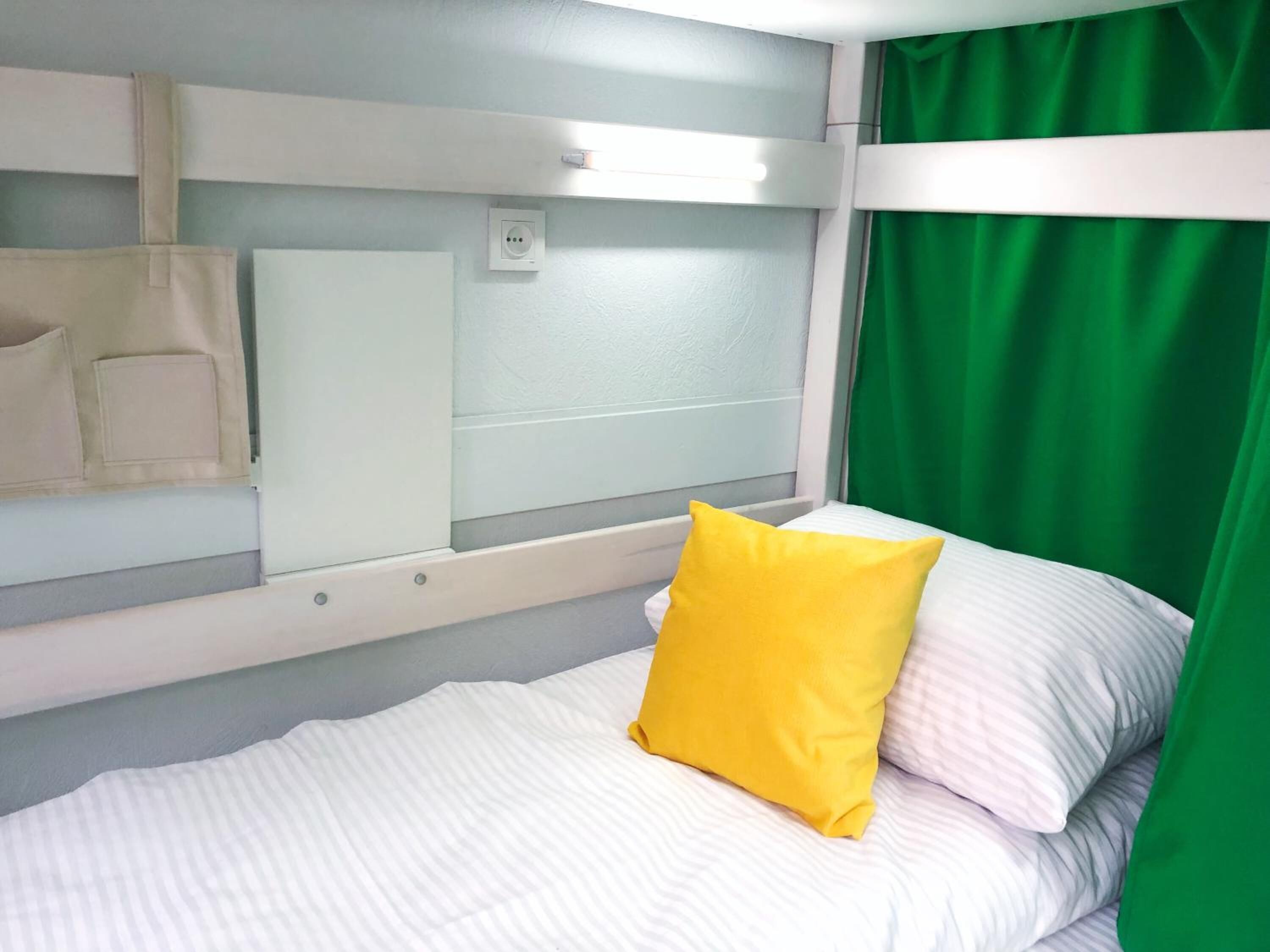 A bed with white bedding and a yellow pillow, a white bedside table, a green curtain, and a white wall with a power outlet.