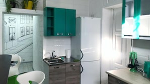 Une cuisine moderne avec des armoires blanches, un évier encastré et un réfrigérateur. On y trouve des plantes en pot sur le plan de travail et une fenêtre donnant sur la rue.