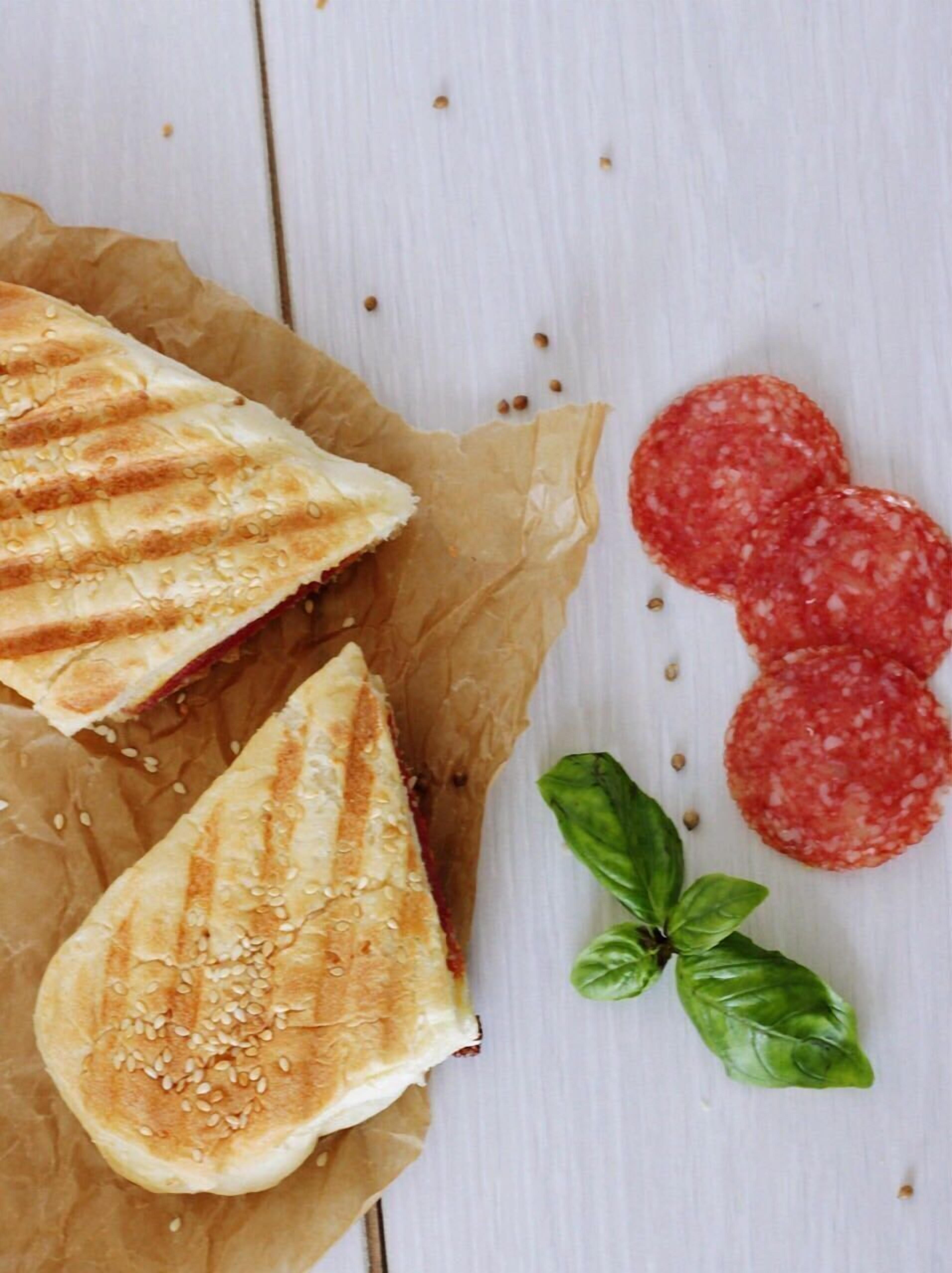 A sandwich with sesame seeds on a piece of parchment paper, accompanied by slices of salami and a basil leaf.