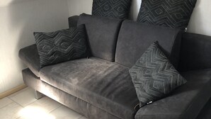 Un sofá gris con cuatro cojines estampados frente a una pared blanca, ubicado en una esquina con piso de baldosas.