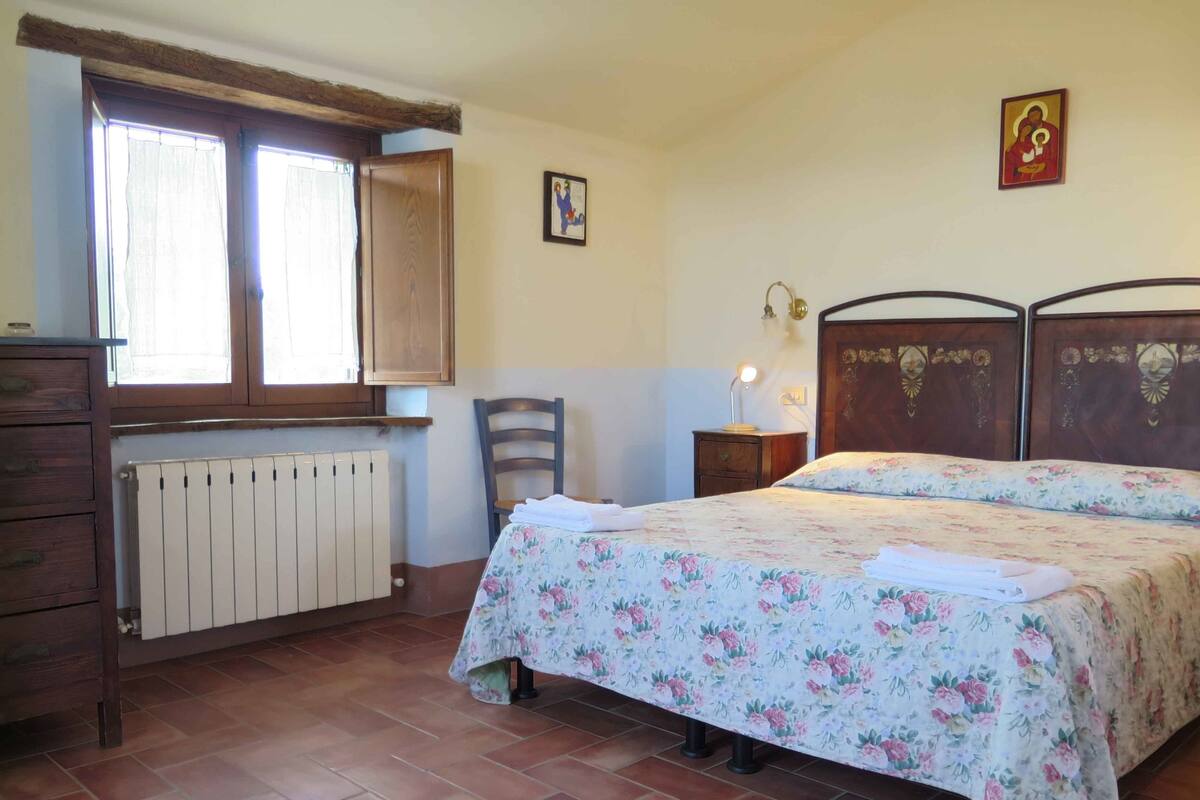Une chambre avec un lit en bois, un radiateur, une fenêtre avec des rideaux, une chaise et une petite table de chevet avec une lampe.