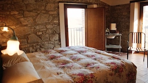 A bedroom with a stone wall, a bed with a floral bedspread, a desk with a chair, and a window with a view. - Agriturismo Santa Maria (San Venanzo)
