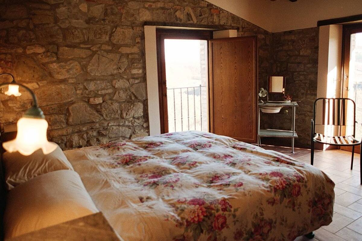 Une chambre avec un mur en pierre, un lit recouvert d’une couvre-lit à motifs floraux, un bureau avec une chaise et une fenêtre donnant sur un paysage agréable.