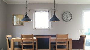 A dining area with a wooden table and chairs, two pendant lights, a wall clock, and a window with natural light.
