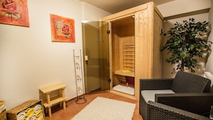 Et sauna-rum med en trædør, en lille træbænk, en hvid måtte, en fletstol og en potteplante.