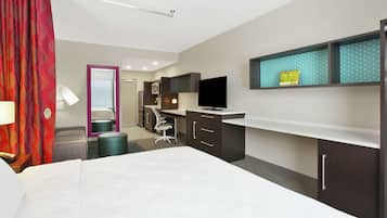 A hotel room with a bed, desk, chair, TV, and a small shelf with a calendar.