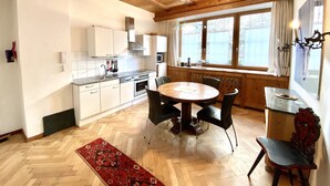 A kitchen with white cabinets, a wooden dining table with chairs, a red patterned rug, and a window with curtains.