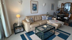 Un salon avec un canapé beige, une table basse en verre et un tapis bleu et beige. On aperçoit une lampe sur une table d’appoint, et un chien est visible au fond.