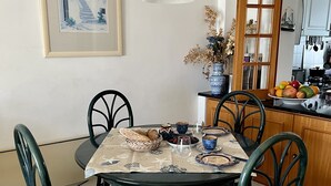 Un coin repas avec une table ronde, des chaises, un tableau encadré au mur, un luminaire suspendu et un espace cuisine visible à travers une porte ouverte.