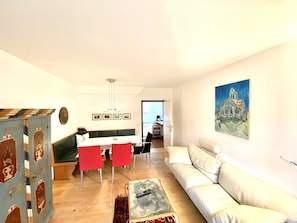 A living room with a white sofa, a dining table with red chairs, a coffee table, and a painting on the wall.
