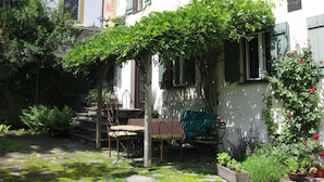 En trädgård med ett träbord och stolar, en pergola med klätterväxter och en vit byggnad med fönster och fönsterluckor.