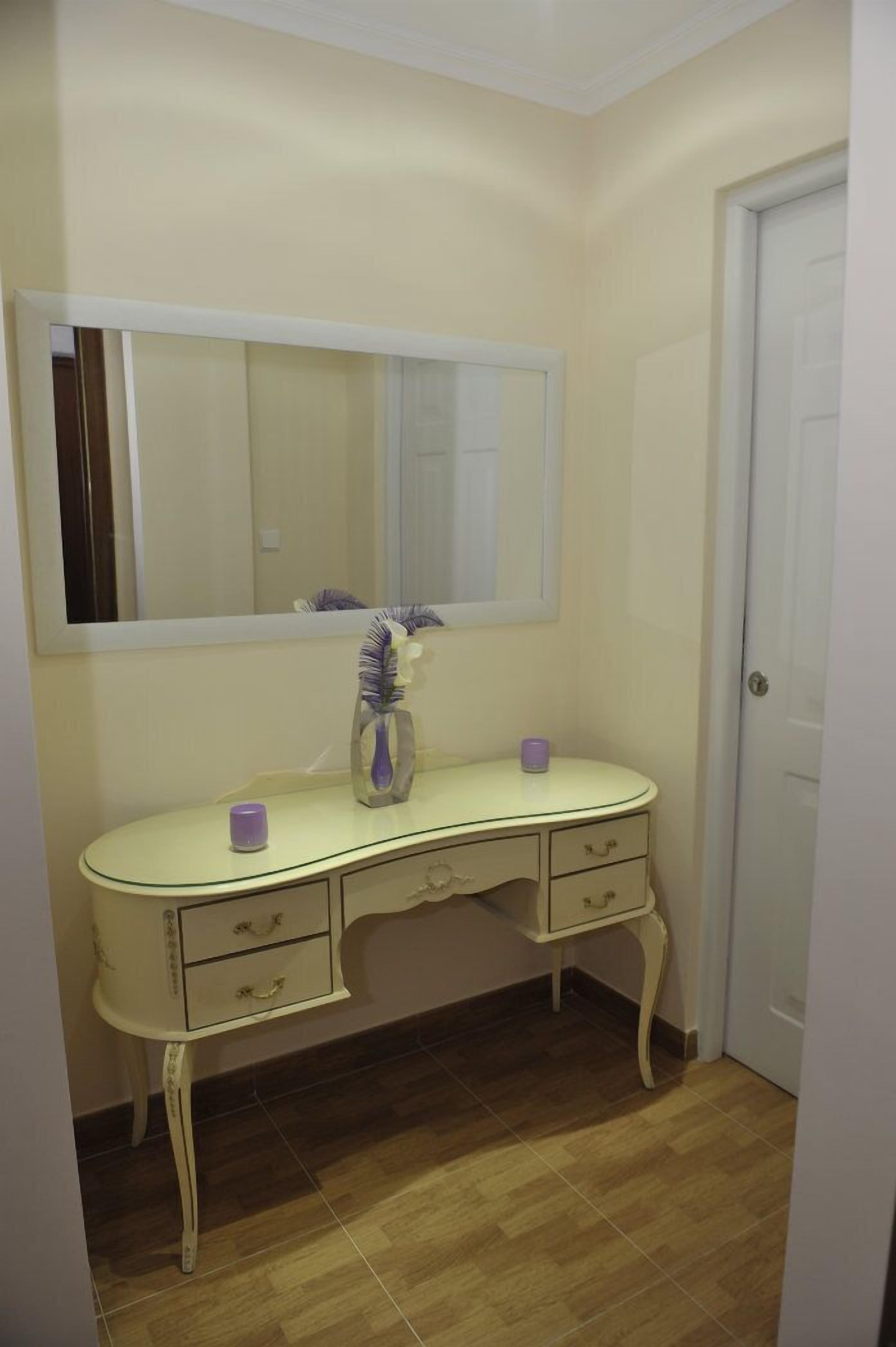 A small, cream-colored vanity with a mirror above it, a vase with a purple candle, and a door to the right.