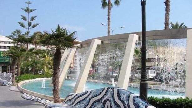 Zona piscina con una poltrona da relax a motivi a mosaico ondulati, palme e cielo sereno.