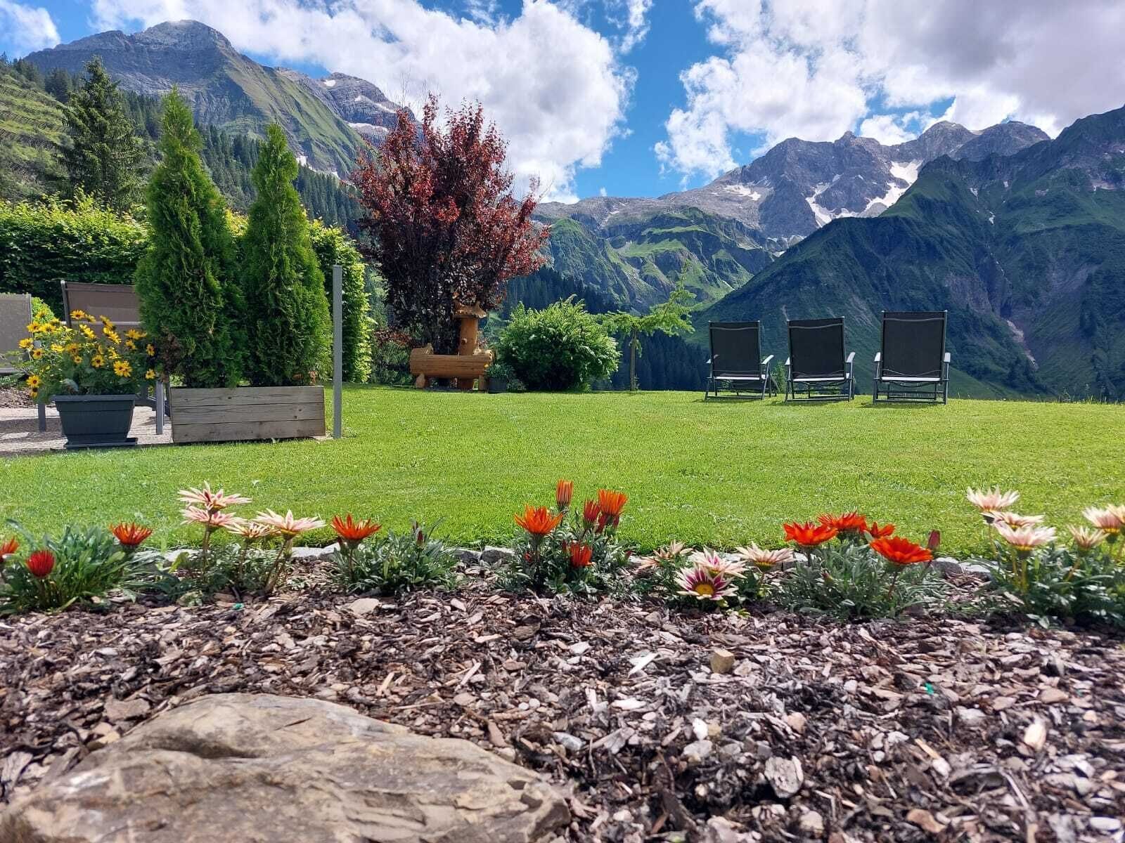 A grassy outdoor area with a mountain backdrop, a wooden sculpture, and three chairs.