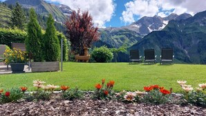 A grassy outdoor area with a mountain backdrop, a wooden sculpture, and three chairs.