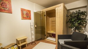 A sauna room with a wooden door, a small wooden bench, a white rug, a wicker chair, and a potted plant.