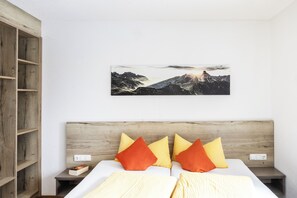 A bedroom with a wooden headboard, a bed with yellow and orange pillows, a nightstand, and a wall-mounted picture of a mountain landscape.