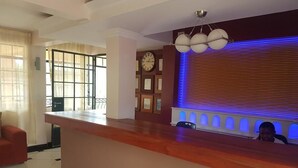 A hotel reception area with a wooden reception desk, a clock, framed pictures on the wall, and a blue-lit wall feature. - Hôtel Le Premier (Kisii)