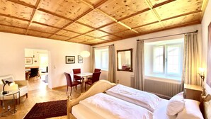 Une pièce avec un plafond en bois, un lit recouvert de draps blancs, une table à manger avec des chaises et une fenêtre avec des rideaux.