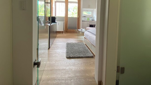 A modern interior with a welcome mat, a kitchen area, and a bedroom with a bed and a window. - Tegernsee - Great, central apartment (Tegernsee)