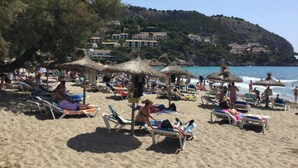 A beach with sun loungers and umbrellas, a clear sky, and buildings in the background. - Dream location in Costa Canyamel (Canyamel)