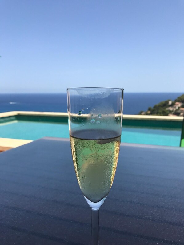 A glass of white wine on a table with a pool and ocean view in the background.