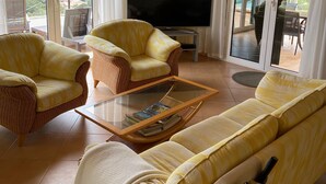 A living room with yellow-striped sofas, a glass coffee table, and wicker chairs. A flat-screen TV is mounted on the wall, and there is a sliding glass door leading to an outdoor area. - Dream location in Costa Canyamel (Canyamel)