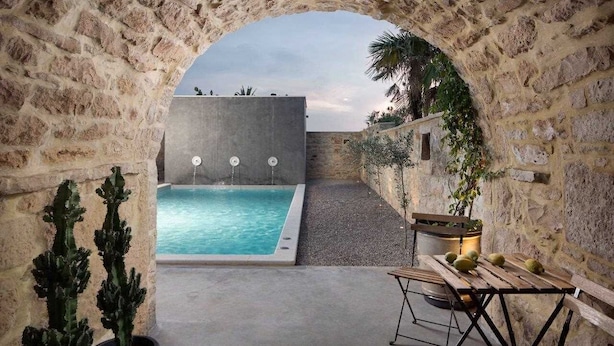 An outdoor pool area with a stone archway, a concrete wall, and a wooden bench with cushions.