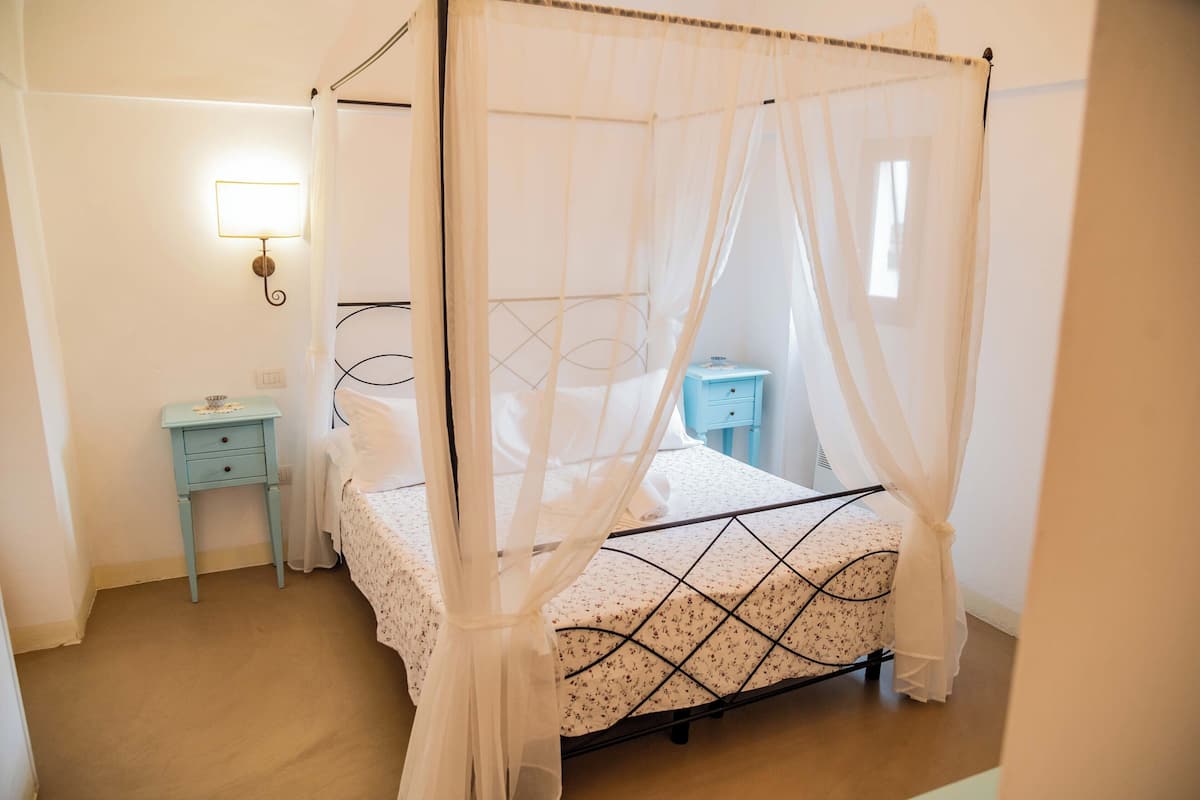 A bedroom with a four-poster bed, a nightstand, a wall-mounted lamp, and a window.