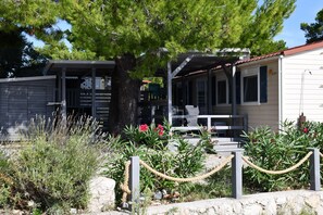 A building with a covered porch, a tree, and a garden with various plants and flowers. - Holiday house / mobile home in the 1. Line seafront, 34 m² + 24 m² terrace (Pirovac)