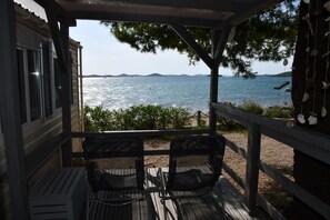 A covered outdoor seating area with two chairs and a table, overlooking a body of water with distant landforms. - Holiday house / mobile home in the 1. Line seafront, 34 m² + 24 m² terrace (Pirovac)