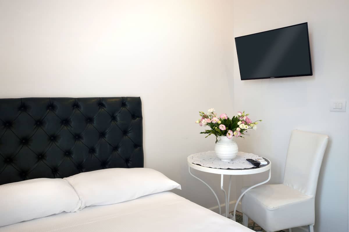 A hotel room with a bed, a white chair, a small round table with a vase of flowers, and a flat-screen TV mounted on the wall.