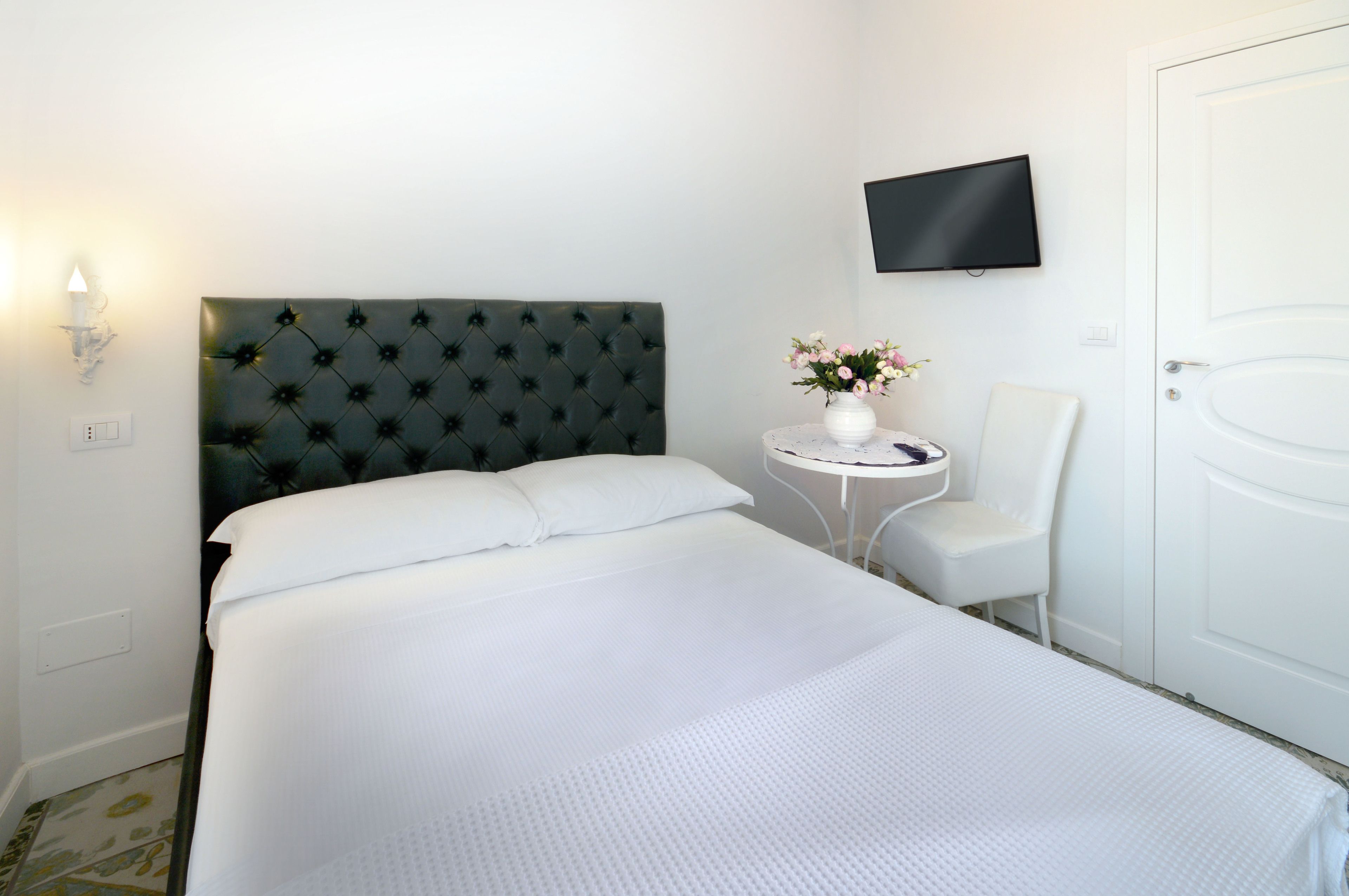 Una camera d'albergo con un letto grande, un tavolino con un vaso di fiori, una sedia bianca, una TV a schermo piatto montata a parete e una porta bianca.