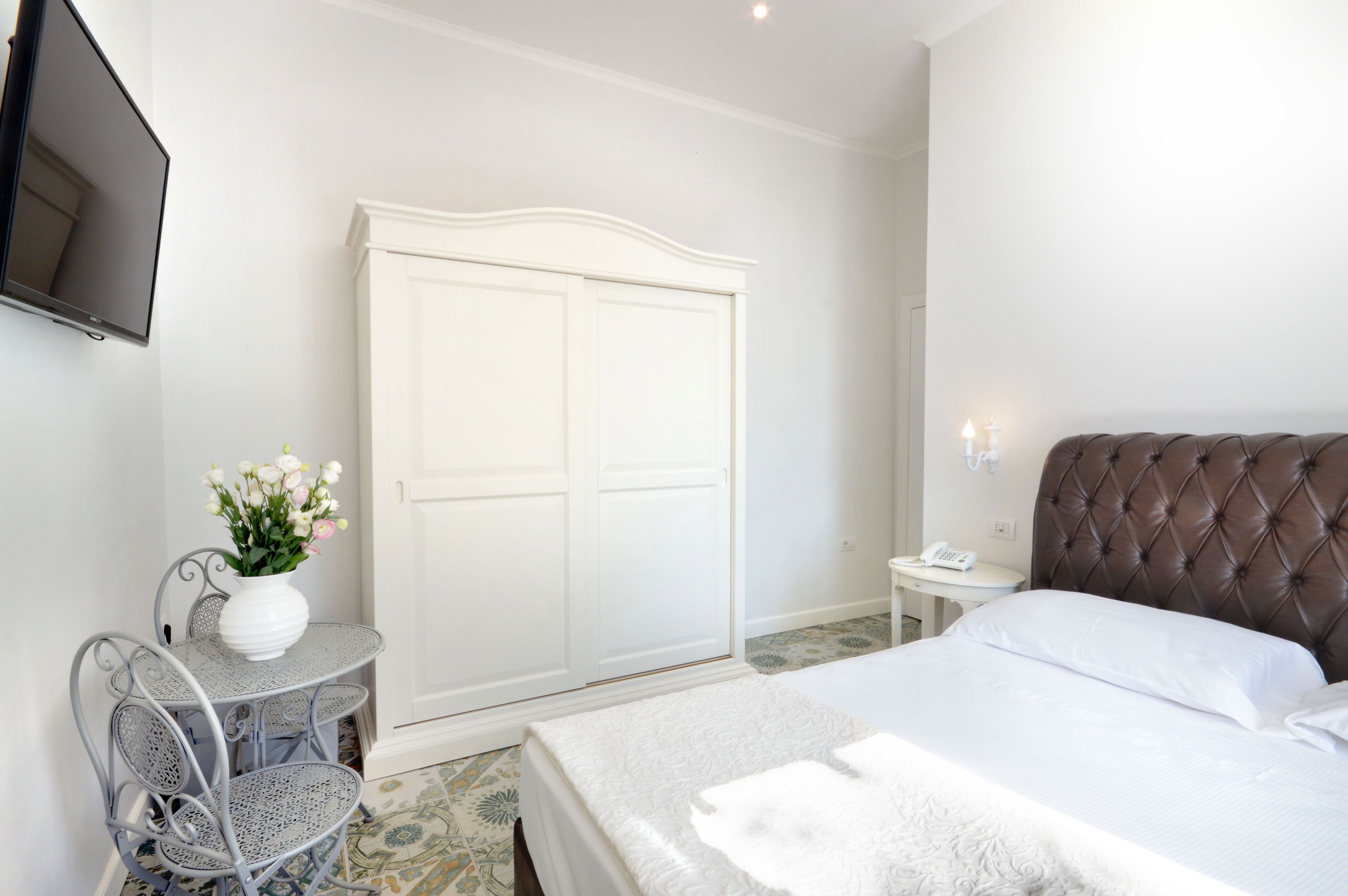 A hotel room with a bed, a white wardrobe, a small table with a vase of flowers, and a television mounted on the wall.
