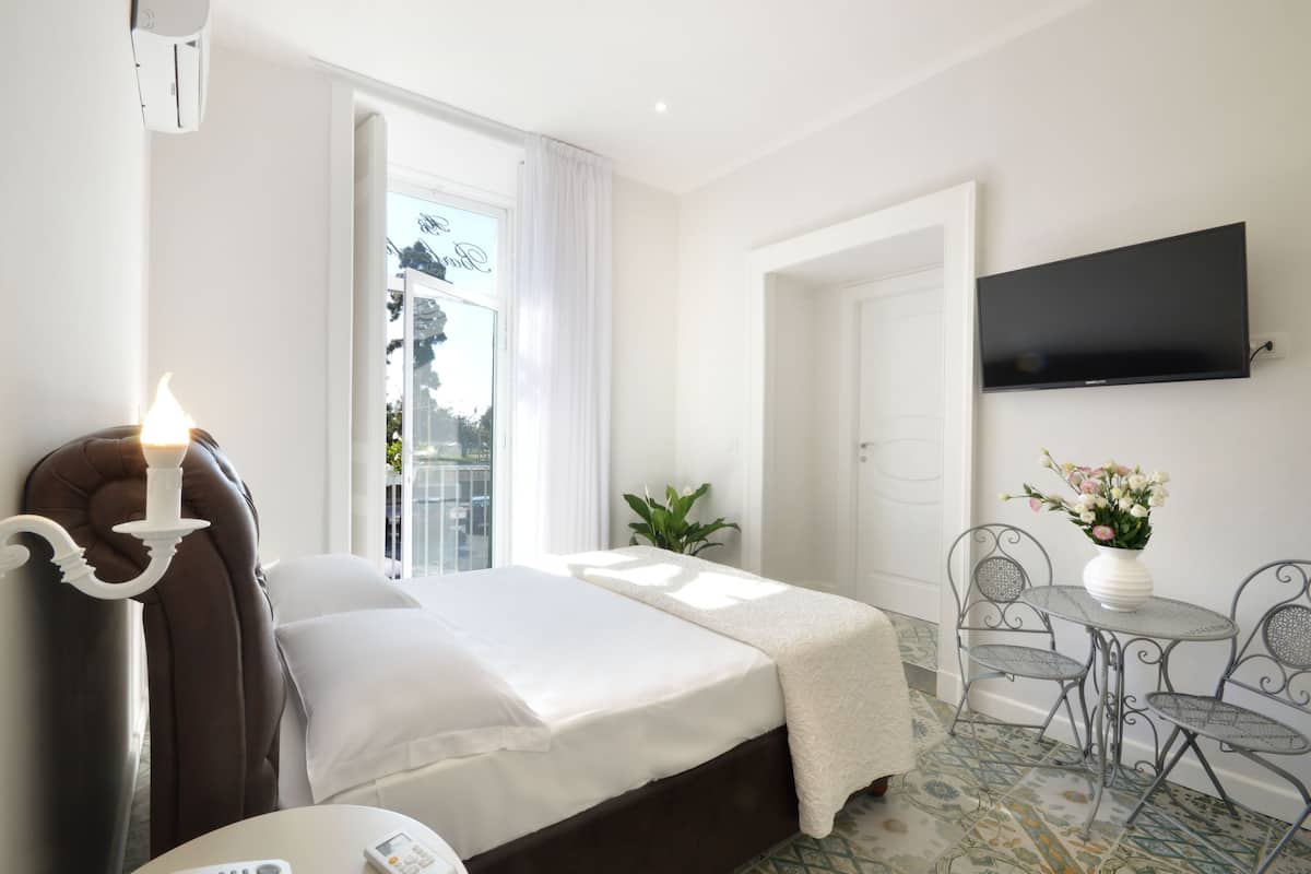 A hotel room with a bed, a TV mounted on the wall, a small table with chairs, and a window with curtains.