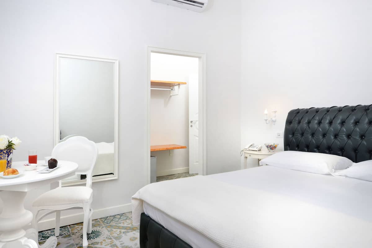 A hotel room with a bed, a bedside table, a chair, a small table with a vase and a muffin, and a wardrobe with a mirror.