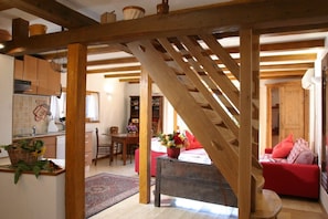Una scala in legno conduce a una zona giorno con un divano rosso, un tavolo da pranzo e una cucina con mobili in legno e un cesto appeso.