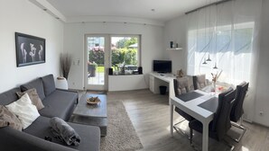 Un saló modern amb un sofà gris, una taula de centre i una zona de menjador amb taula i cadires blanques. Hi ha un televisor sobre un moble baix i una finestra gran amb cortines.