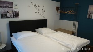 Ein ordentlich bezogenes Bett in einem Hotelzimmer, mit einem schwarzen Kopfteil, einem kleinen Holztischchen und einem an der Wand befestigten Wäscheständer.
