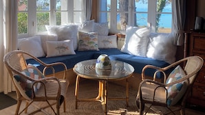 A cozy room with a blue sofa, wicker chairs, a glass coffee table, and large windows offering a view of trees and a body of water. - Vacation home "Bord de Mer" - Exceptional sea view (Petit-Caux)