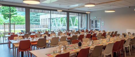 A spacious dining hall with long tables set for a meal, large windows offering a view of the outdoors, and pendant lighting.