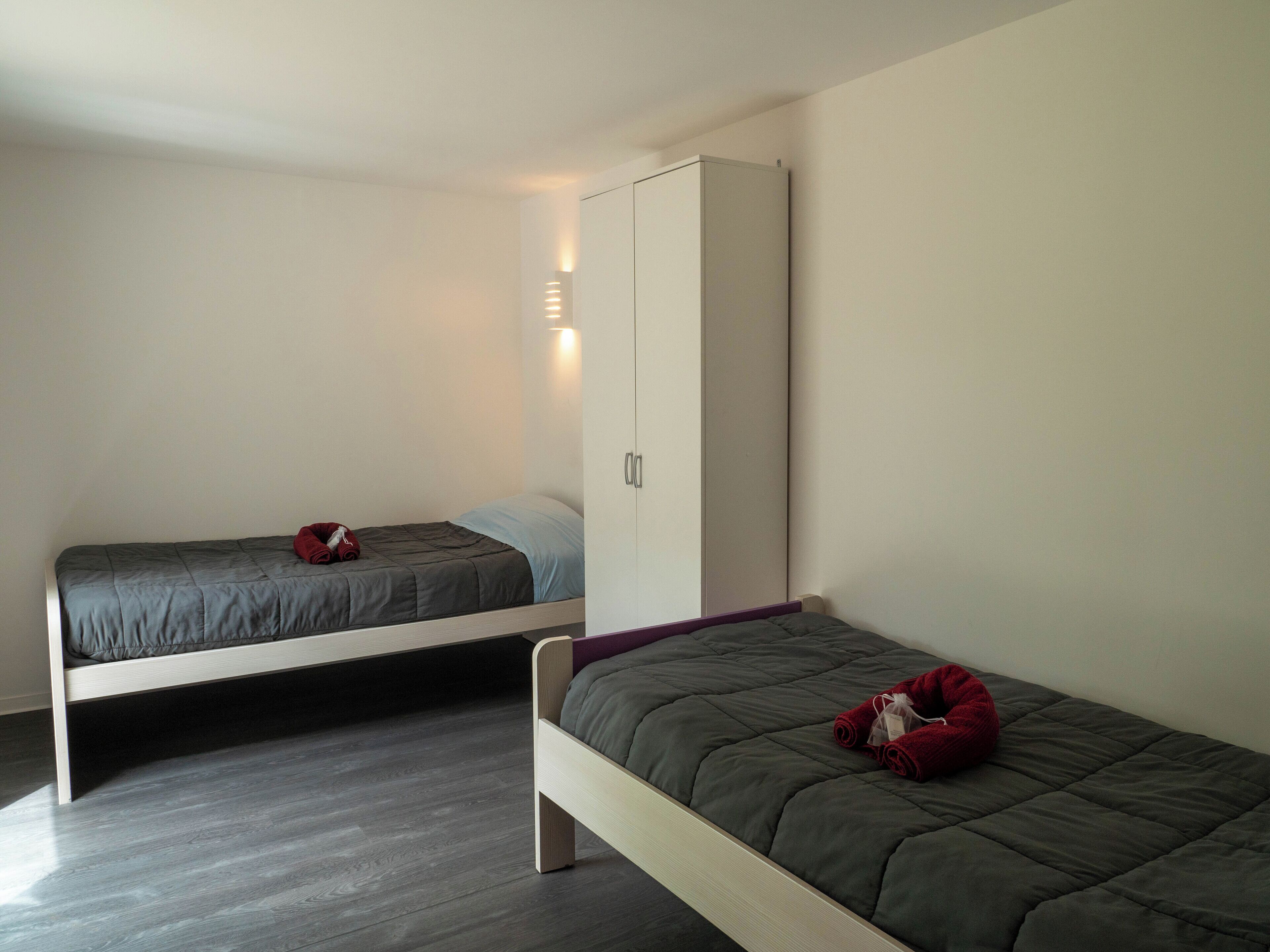 Two single beds with grey bedding, a white wardrobe, and a wooden floor.