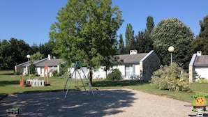 Une aire de jeux avec des balançoires et une petite structure d’escalade, entourée d’une zone recouverte de gravier et parsemée de quelques touffes d’herbe.